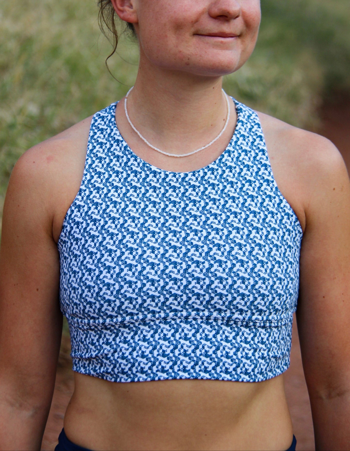 A woman outdoors wearing a blue and white patterned athletic crop top, with sunglasses on her head and a thin silver necklace.
