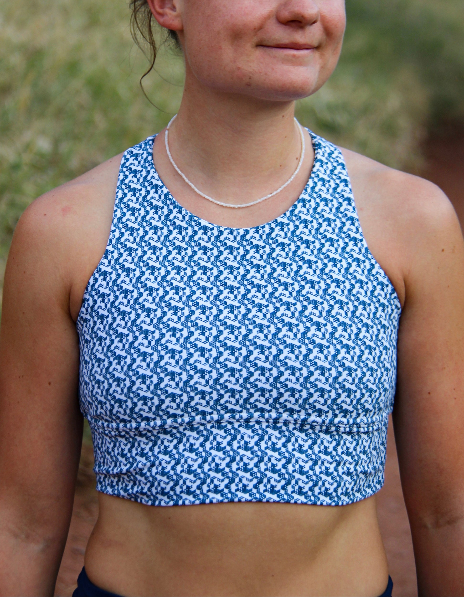 A woman outdoors wearing a blue and white patterned athletic crop top, with sunglasses on her head and a thin silver necklace.