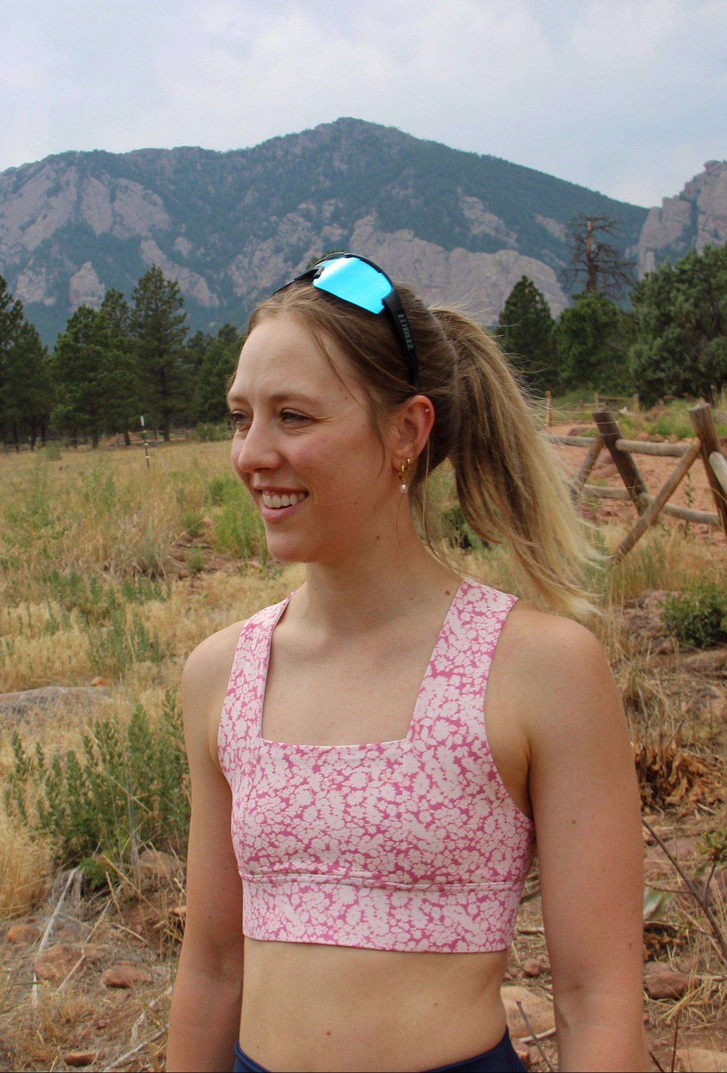A woman wearing a pink and white patterned sports bra stands outdoors in a grassy mountain landscape.