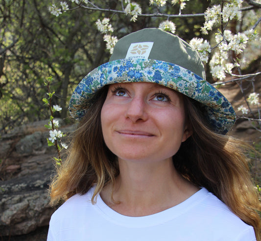 Woman wearing a white shirt and floral hat outdoors with trees and flowers in the background